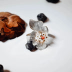 Front view of a silver flower ring with open cupped petals and a pearl and coral centre cluster with gold accent beads, surrounded by a pine cone and dark stones on a white surface.