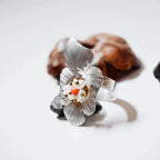 Side view of a silver flower ring with softly curved textured petals and a raised pearl and coral gemstone centre, resting on dark pebbles with a pine cone in the background.