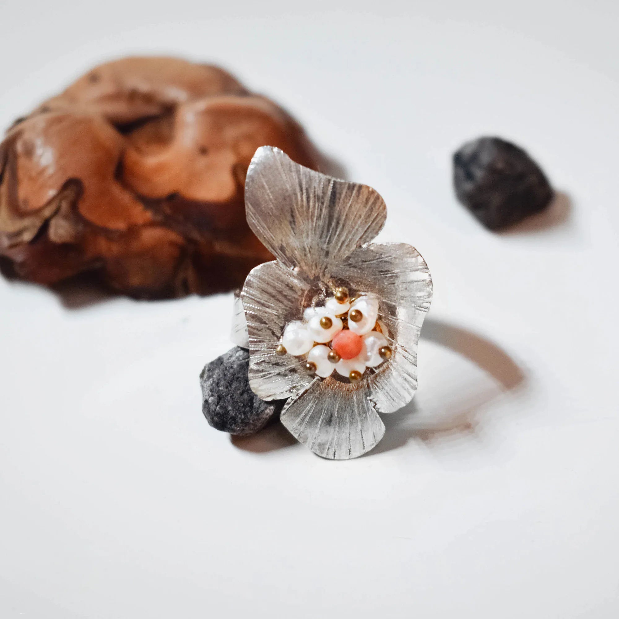 Silver flower ring with four cupped textured petals and a cluster of white seed pearls and a pink coral bead at the centre, leaning against a pine cone and dark pebble on a white surface. Main image