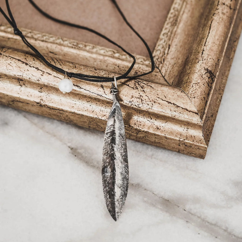 Oxidised silver olive leaf pendant on a black cord with a single white freshwater pearl accent knotted to the side, displayed against a vintage gold frame on marble.