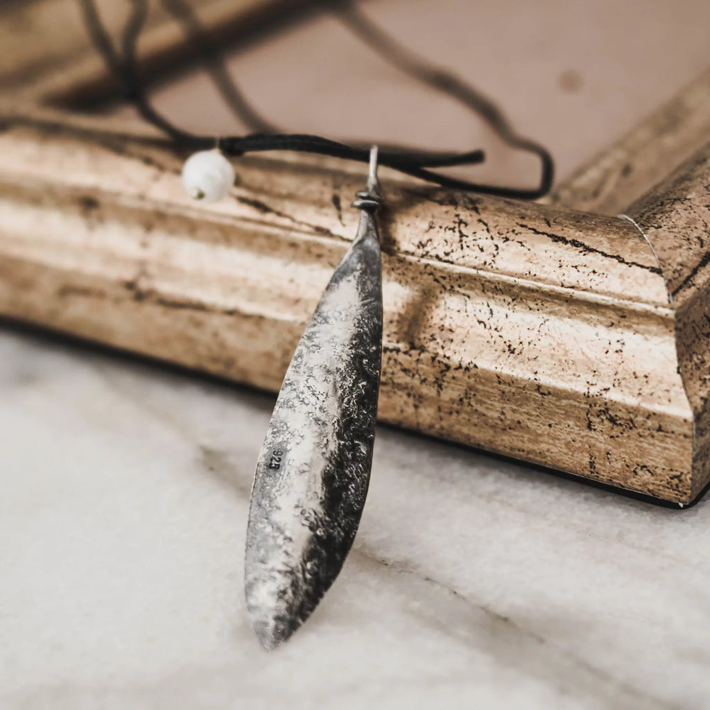 Reverse side of an oxidised silver olive leaf pendant showing the 925 hallmark stamp and textured surface, displayed against a vintage gold frame on marble.