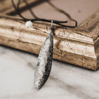 Reverse side of an oxidised silver olive leaf pendant showing the 925 hallmark stamp and textured surface, displayed against a vintage gold frame on marble.