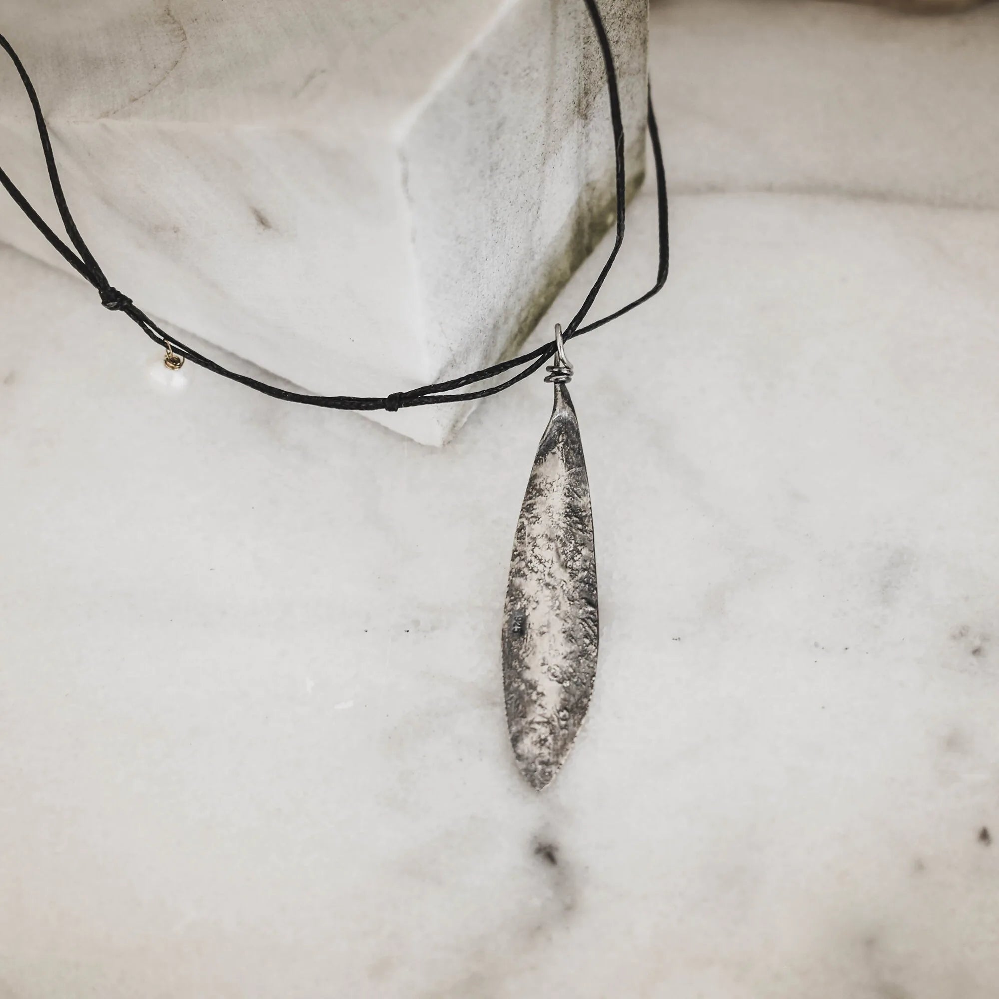 Long slender oxidised silver olive leaf pendant with a deeply textured surface, hanging on a black cord against a white marble surface. Main image