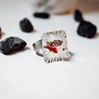 Front view of a square silver ring with radiating texture and a coral, amethyst and pearl gemstone cluster at its centre, surrounded by dark rough stones on a white surface.
