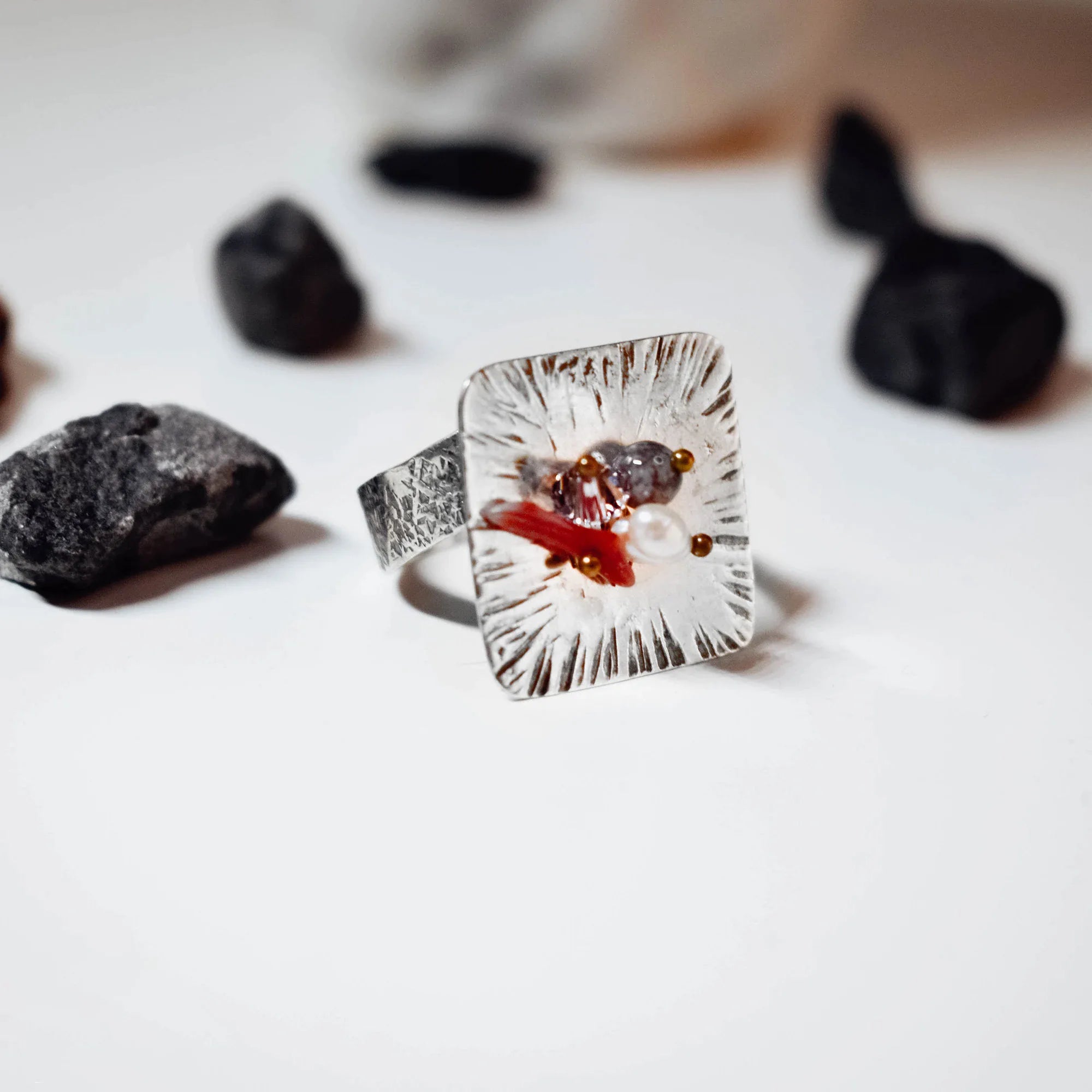 Front view of a square silver ring with radiating texture and a coral, amethyst and pearl gemstone cluster at its centre, surrounded by dark rough stones on a white surface.