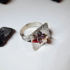 Angled side view of a square silver ring with radiating line texture and a raised gemstone cluster of red, purple and white stones, with a dark pebble beside it on a white surface.