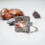 Large square silver ring with rounded corners, deep radiating line texture and a small cluster of pink, purple and white stones with gold beads at the centre, displayed on a white surface with a pine cone and dark stone in the background.