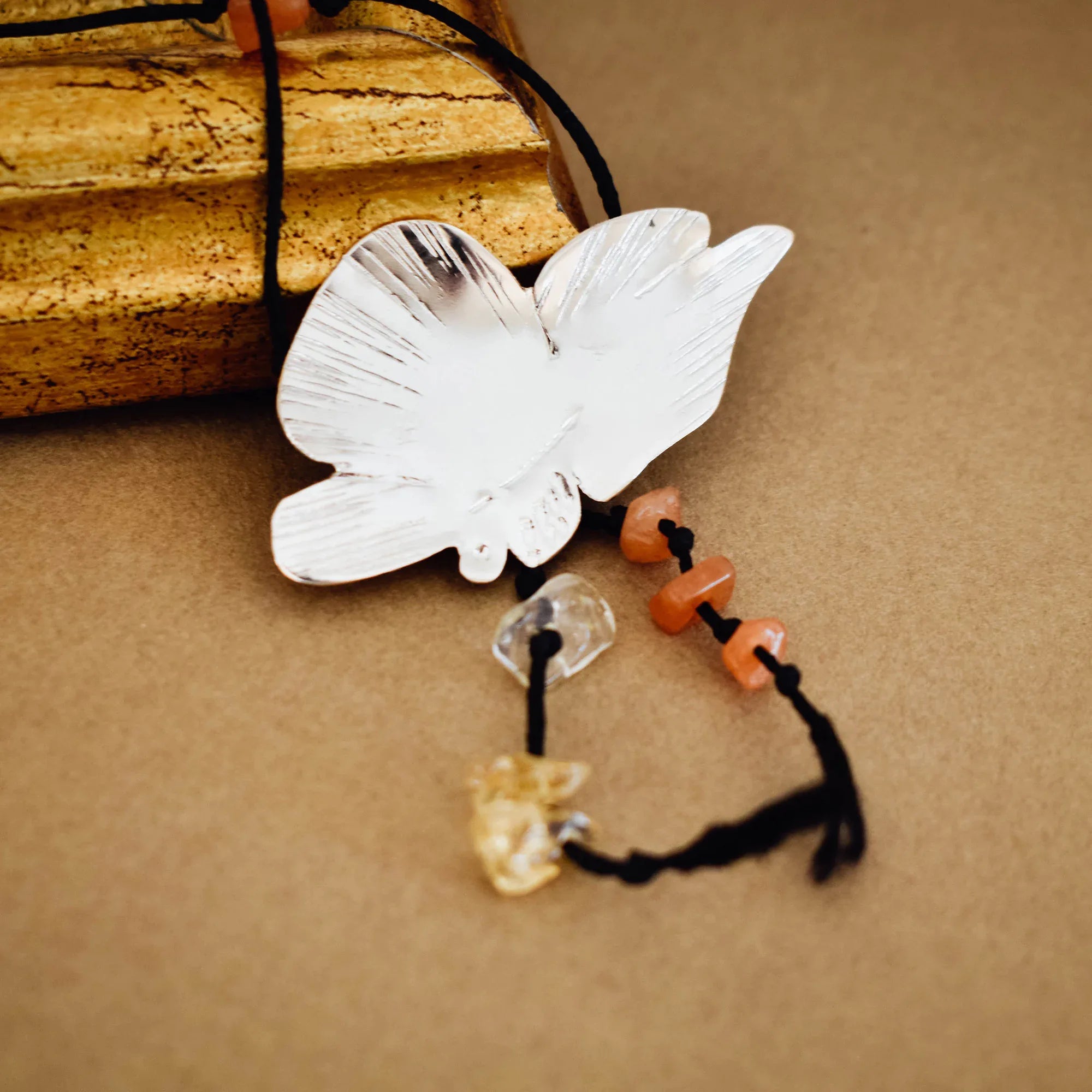 Close-up of a silver butterfly pendant showing the brushed engraved wing detail with orange agate and pale honey crystal chip drops hanging from the body on a black tassel cord.