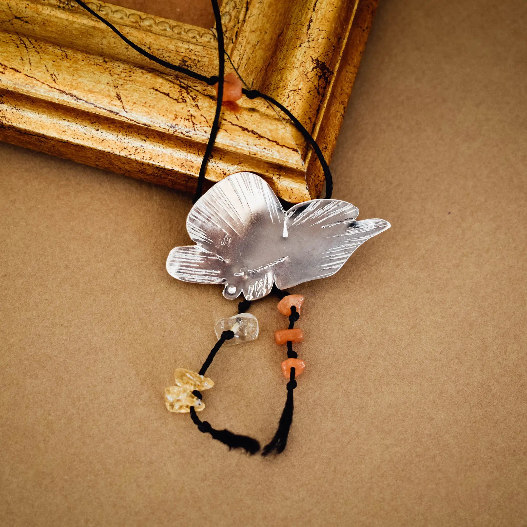 Silver butterfly pendant with engraved wings and orange agate and pale crystal chip drops on a black tassel cord, displayed against a vintage gold frame on a brown surface. Main image