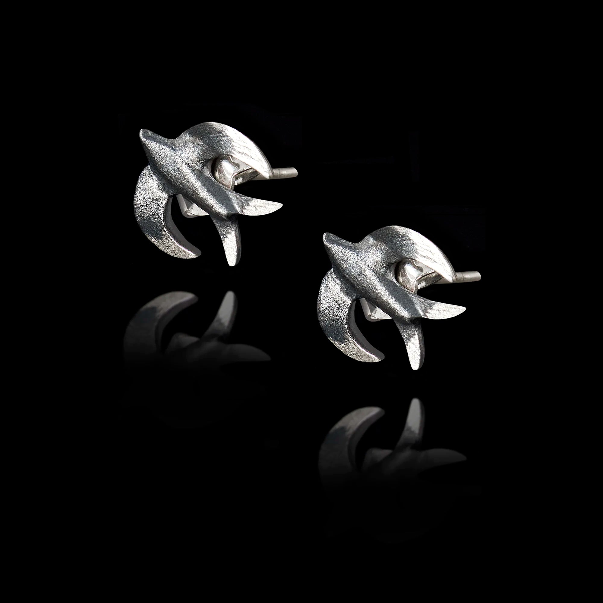 Angled side view of a pair of silver stud earrings shaped like swallows with a dark oxidized finish.