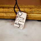 Close-up of a rectangular silver pendant showing the oxidised bark texture with raised polished silver heart, signpost and horizontal bar elements on a black cord against a gold frame.