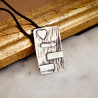 Rectangular silver pendant with deeply scratched bark-like oxidised texture and raised polished heart, signpost and two horizontal bar details, on a black cord against a vintage gold frame.