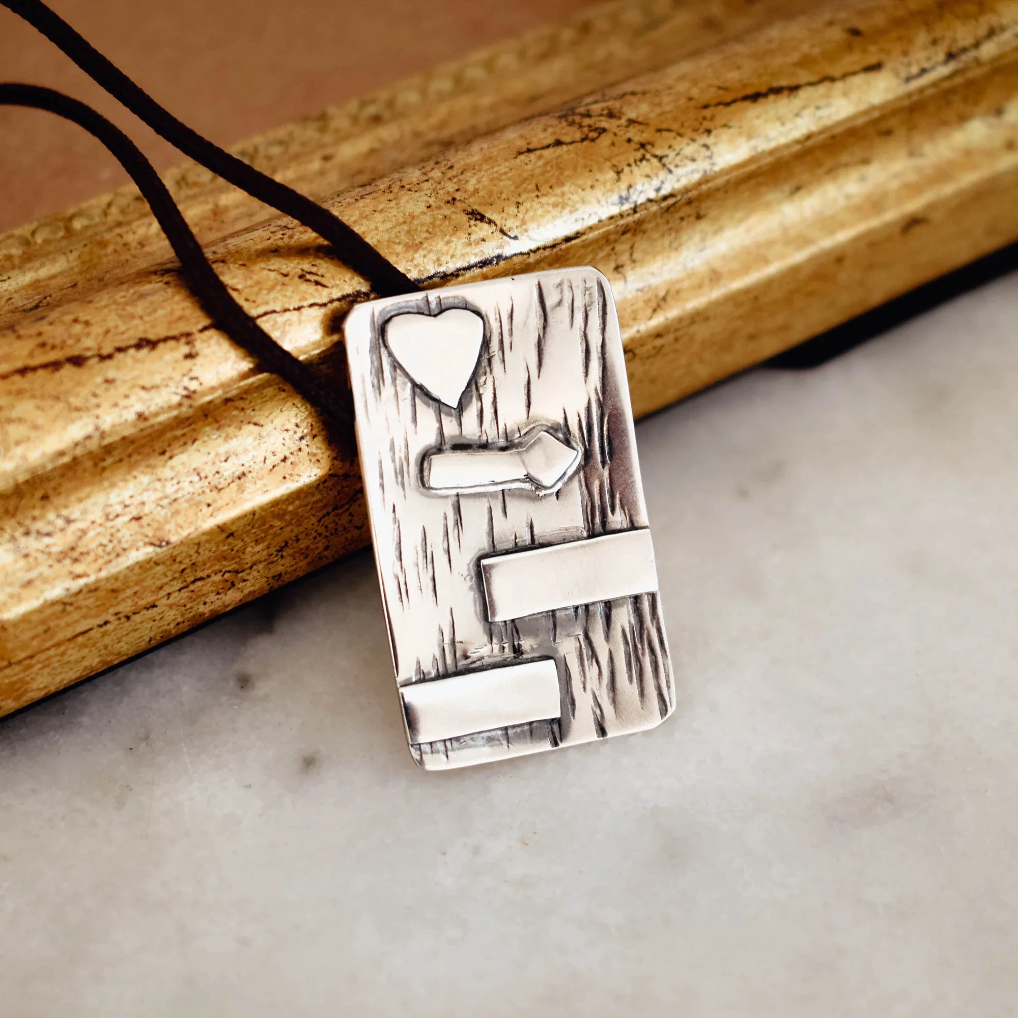 Rectangular silver pendant with deeply scratched bark-like oxidised texture and raised polished heart, signpost and two horizontal bar details, on a black cord against a vintage gold frame. Main image