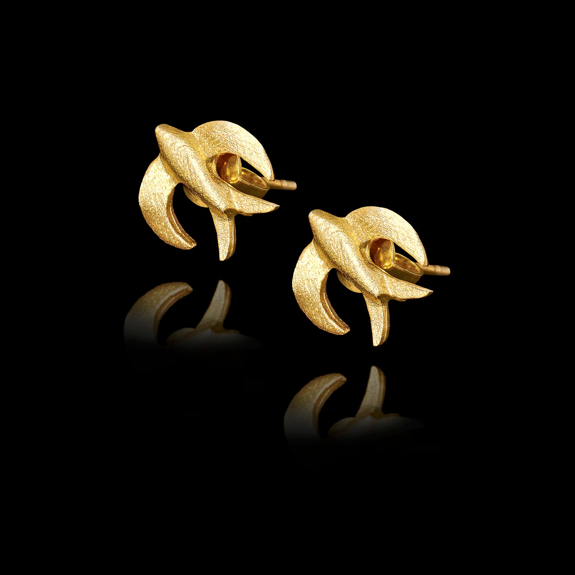 angled side view of  gold stud earrings shaped like swallows in flight on black background.