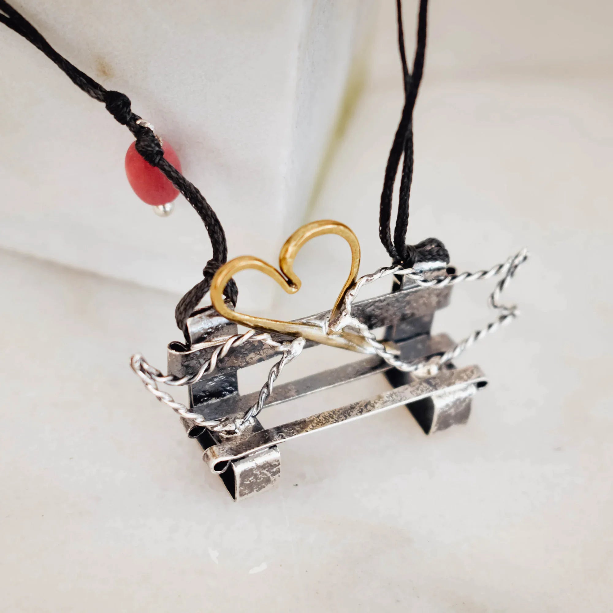 Close-up of an oxidised silver bench pendant showing the gold-plated wire heart detail, twisted silver wire sides and horizontal bar bench structure on a double-strand black cord.