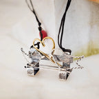 Miniature oxidised silver bench pendant with twisted wire side details and a gold-plated wire heart above, on a double-strand black cord with a red coral bead accent, against a marble surface.