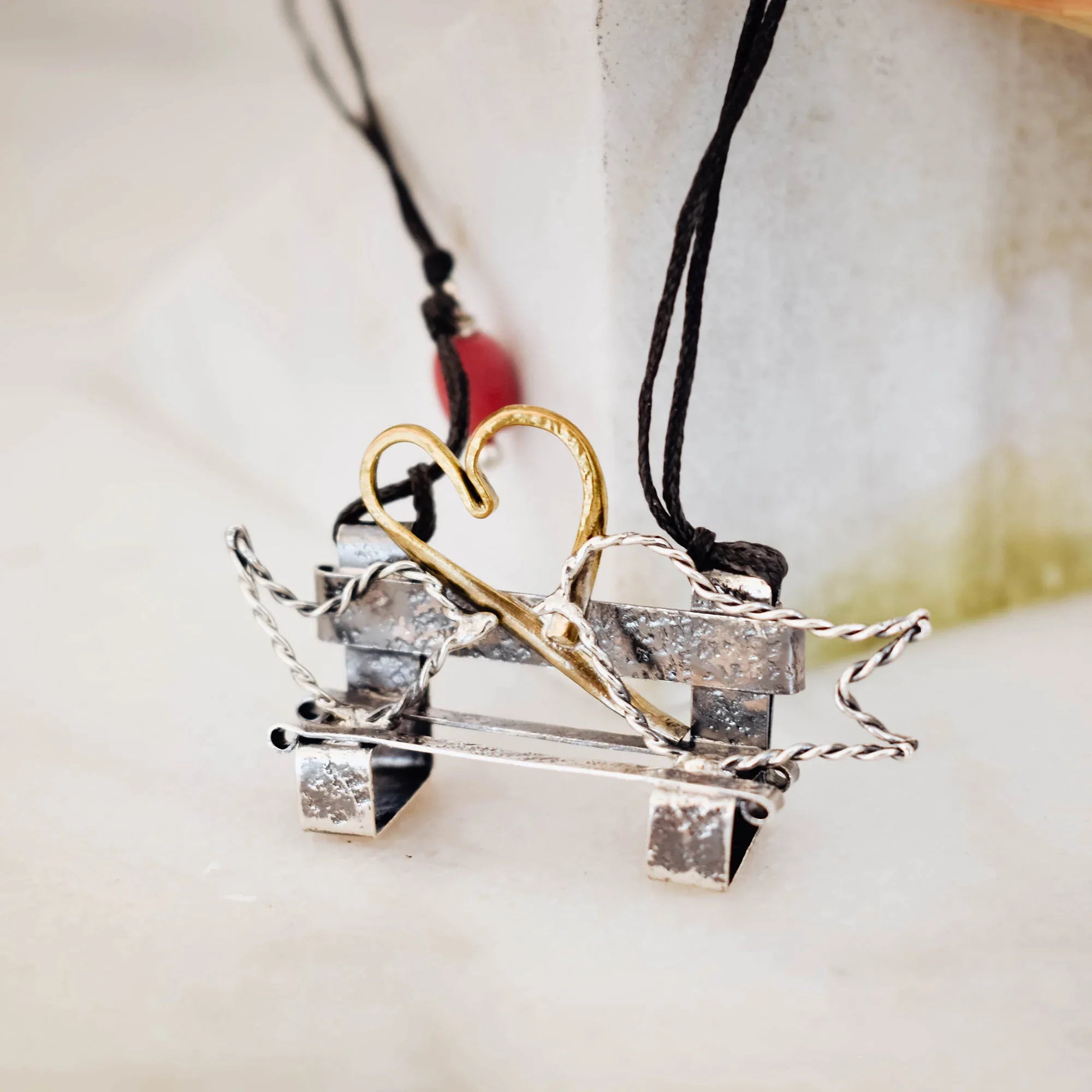 Miniature oxidised silver bench pendant with twisted wire side details and a gold-plated wire heart above, on a double-strand black cord with a red coral bead accent, against a marble surface. Main image