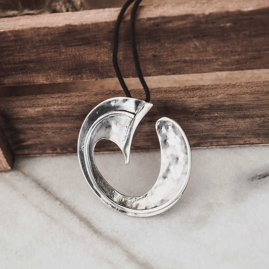 Front view of a large hammered silver wave pendant showing the circular arc form and inner curved detail on a black cord against a wooden surface and marble.