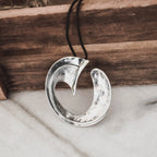 Front view of a large hammered silver wave pendant showing the circular arc form and inner curved detail on a black cord against a wooden surface and marble.