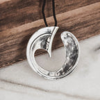 Large hammered silver wave pendant with a bold circular arc and inner curved element on a black cord, displayed against a wooden surface and white marble.