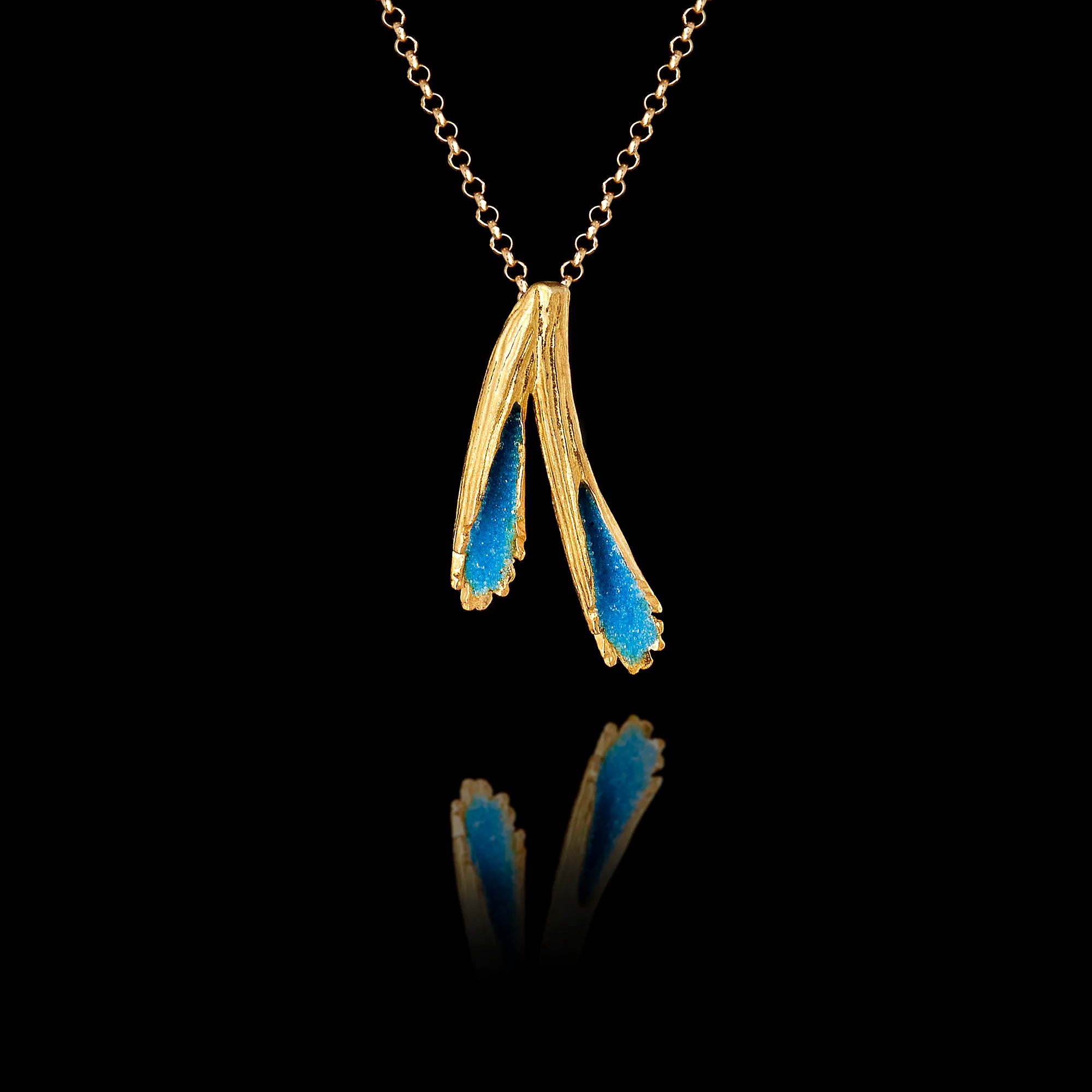 A gold pendant featuring two textured forms filled with vivid blue crushed enamel crystals, hanging on a gold chain against a black background. Main image
