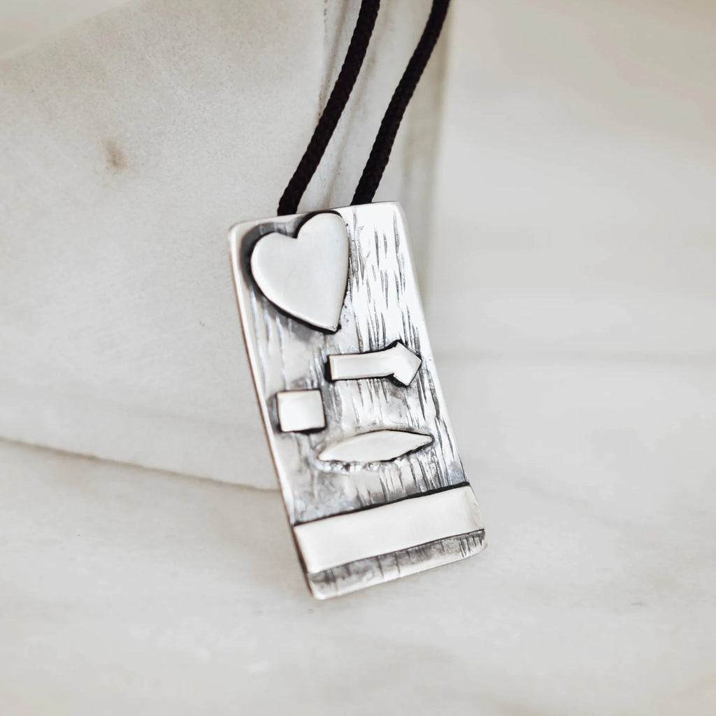 Close-up of a rectangular silver pendant showing the deeply scratched oxidised bark texture with raised polished silver heart, arrow, square, leaf and bar elements on a black cord.