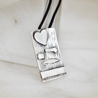 Close-up of a rectangular silver pendant showing the deeply scratched oxidised bark texture with raised polished silver heart, arrow, square, leaf and bar elements on a black cord.