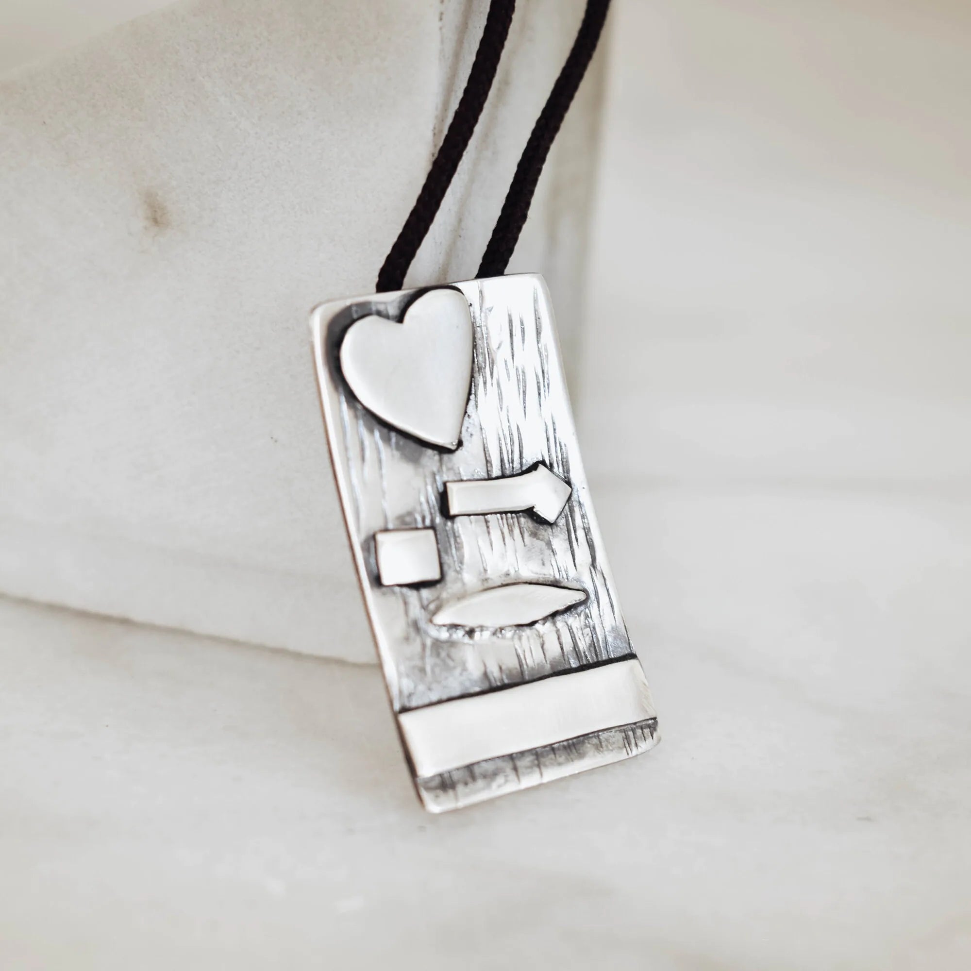 Close-up of a rectangular silver pendant showing the deeply scratched oxidised bark texture with raised polished silver heart, arrow, square, leaf and bar elements on a black cord.