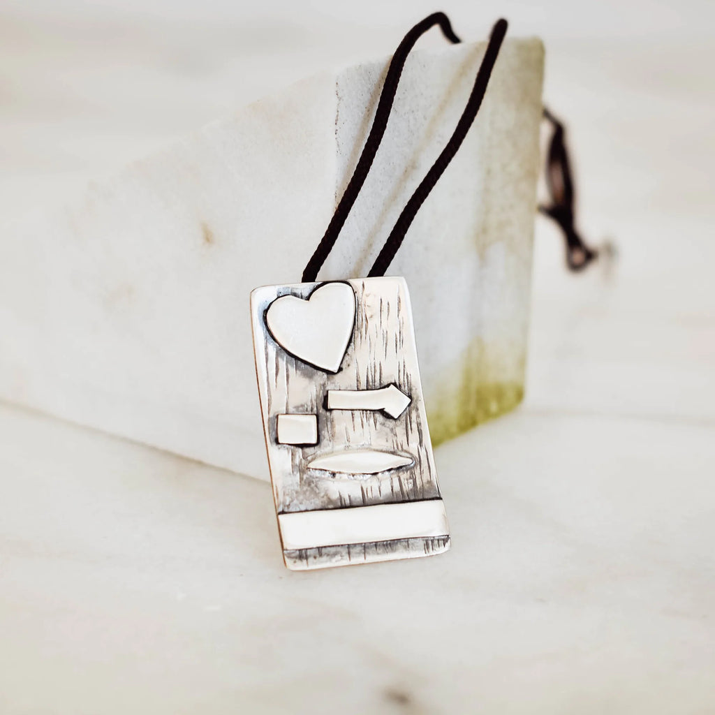 Rectangular silver pendant with bark-textured oxidised surface and raised polished heart, arrow, square, leaf and horizontal bar details on a black cord against a marble surface.