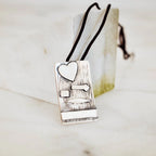 Rectangular silver pendant with bark-textured oxidised surface and raised polished heart, arrow, square, leaf and horizontal bar details on a black cord against a marble surface.