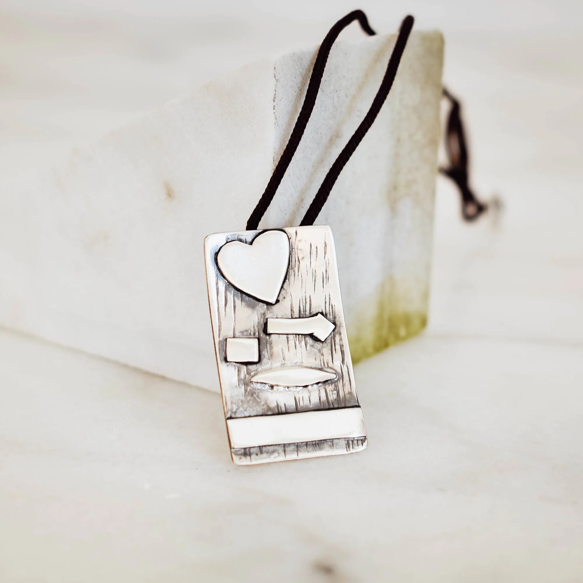 Rectangular silver pendant with bark-textured oxidised surface and raised polished heart, arrow, square, leaf and horizontal bar details on a black cord against a marble surface. Main image