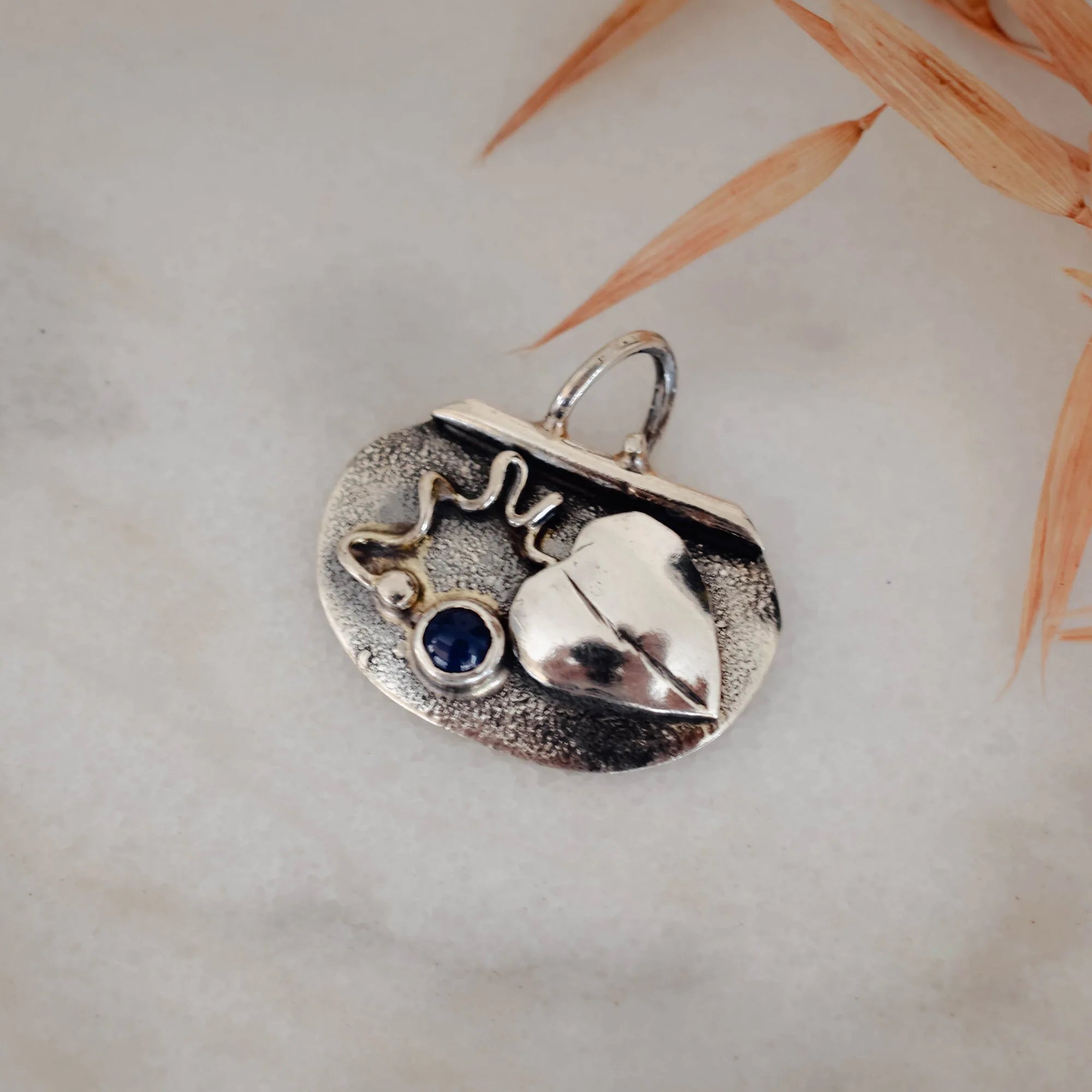 Close-up of a small oval oxidised silver pendant showing the raised polished heart, deep blue lapis lazuli stone, wire scroll and silver granule detail against the dark oxidised background.