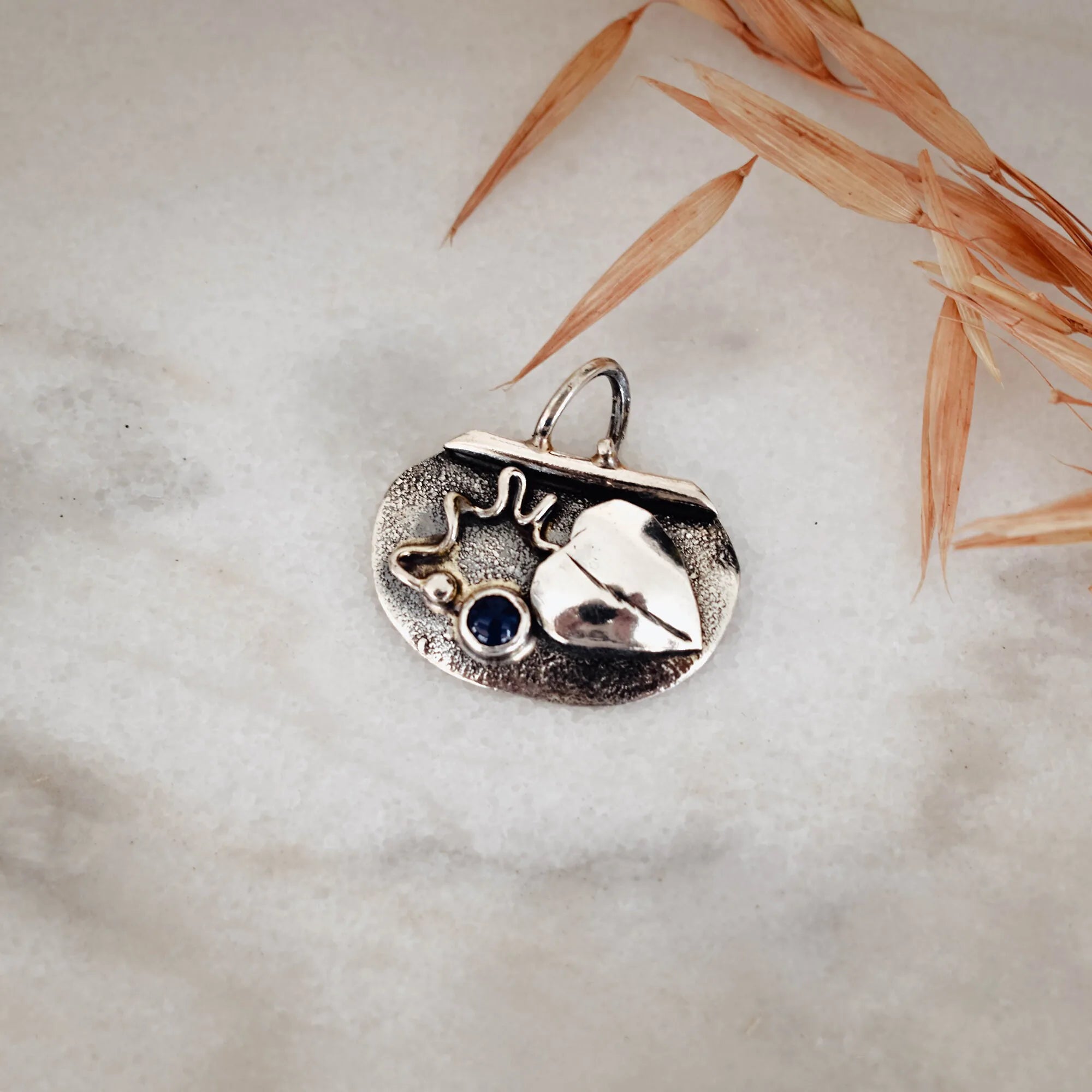 Small oval oxidised silver pendant with a raised polished heart, bezel-set deep blue lapis lazuli cabochon, silver granule and wire scroll detail, displayed on a marble surface with dried grasses. Main image