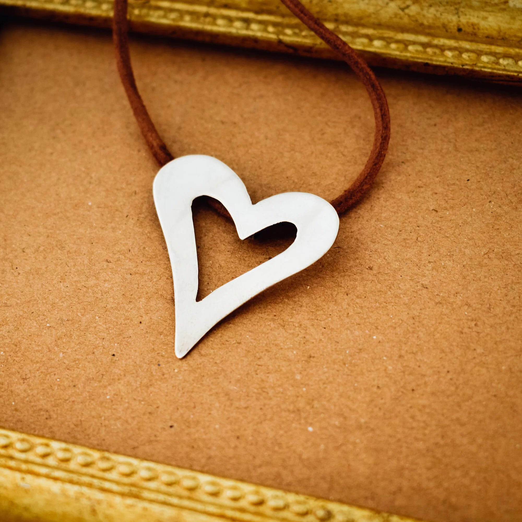 Close-up of a large open heart silver pendant showing the fluid asymmetric outline and brushed silver surface on a brown suede cord.