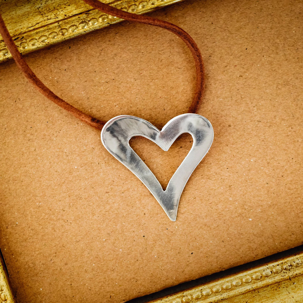 Large open heart silver pendant with brushed surface on a brown suede cord, displayed on a brown surface with a gold vintage frame.