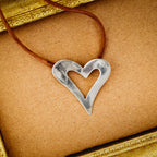 Large open heart silver pendant with brushed surface on a brown suede cord, displayed on a brown surface with a gold vintage frame.