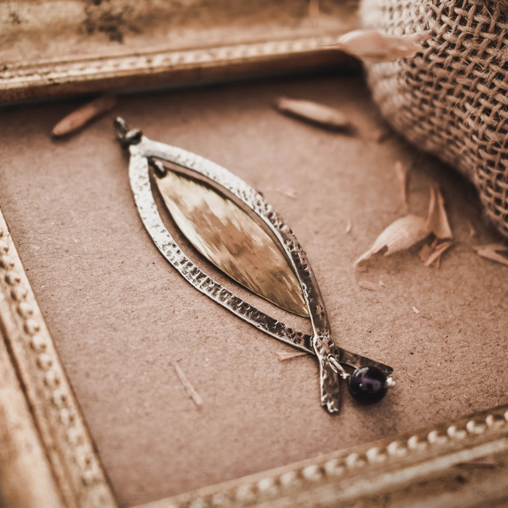 Close-up of a long fish pendant showing the textured oxidised silver frame, smooth gold-plated fish body and forked tail extensions with a small amethyst bead drop on a brown surface.