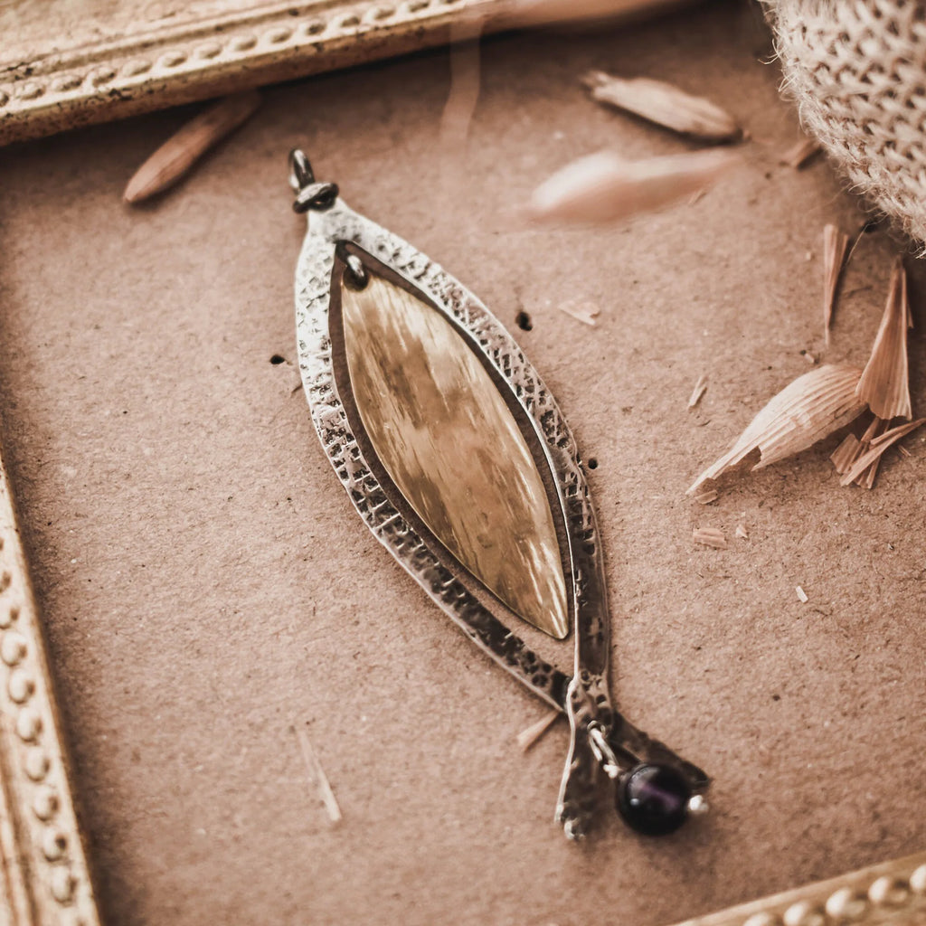 Long fish pendant with a sand-textured oxidised silver outer frame, gold-plated inner body and forked tail with an amethyst bead drop, displayed on a brown surface with a vintage gold frame and dried grasses.