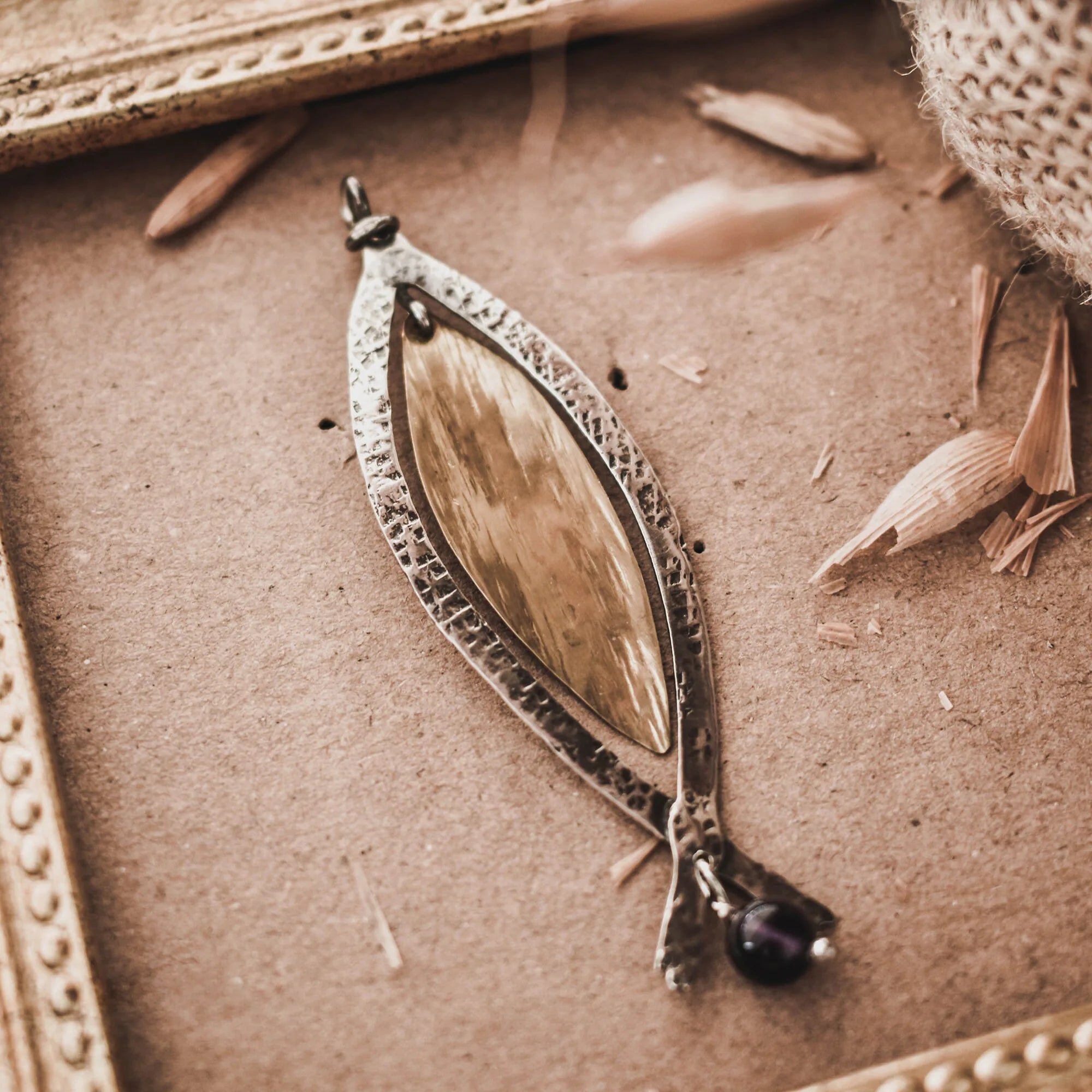 Long fish pendant with a sand-textured oxidised silver outer frame, gold-plated inner body and forked tail with an amethyst bead drop, displayed on a brown surface with a vintage gold frame and dried grasses. Main image
