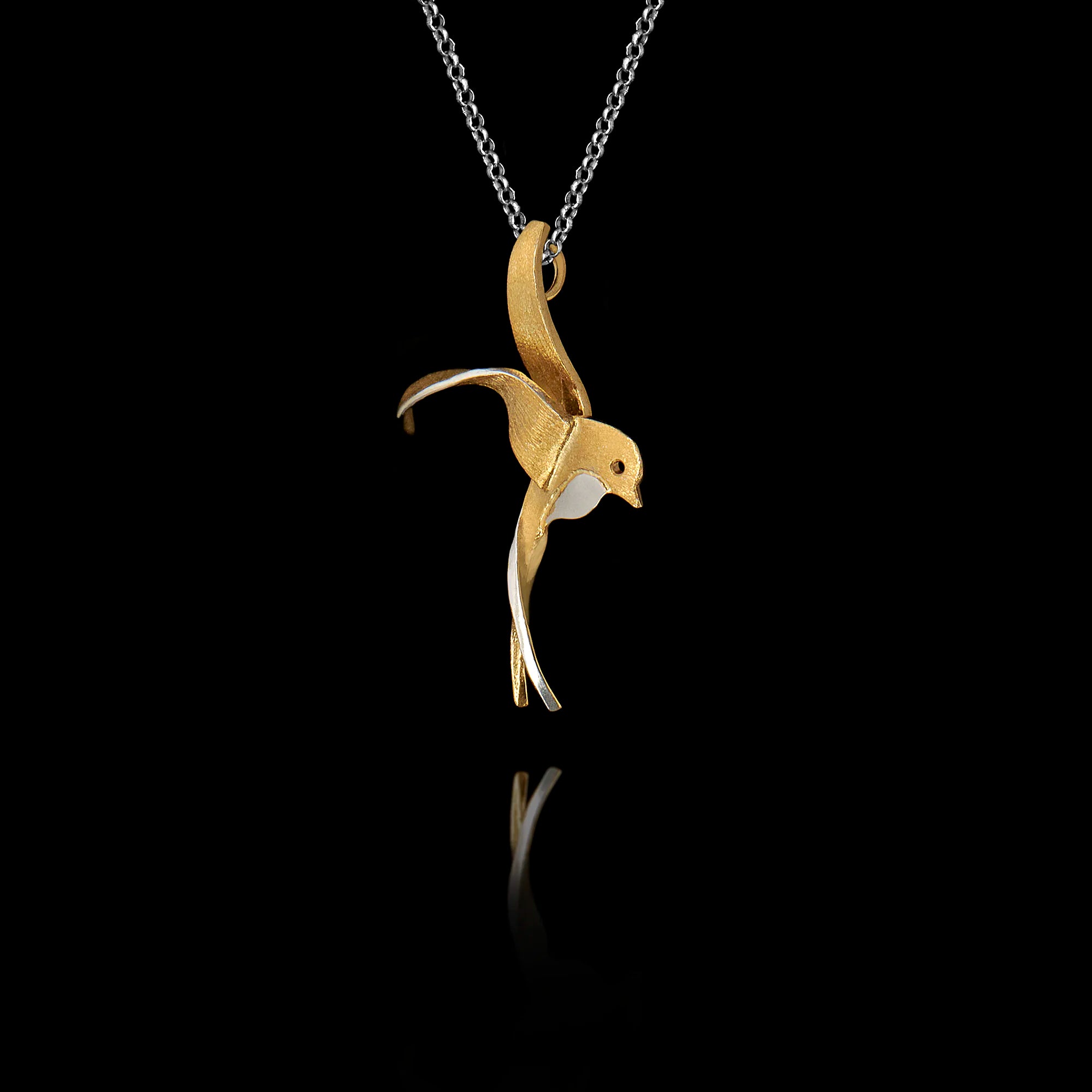 Side angle view of a sterling silver gold-plated swallow pendant with rhodium accent showing the fluid sculpted silhouette of a swallow in flight on a silver chain against a black background.
