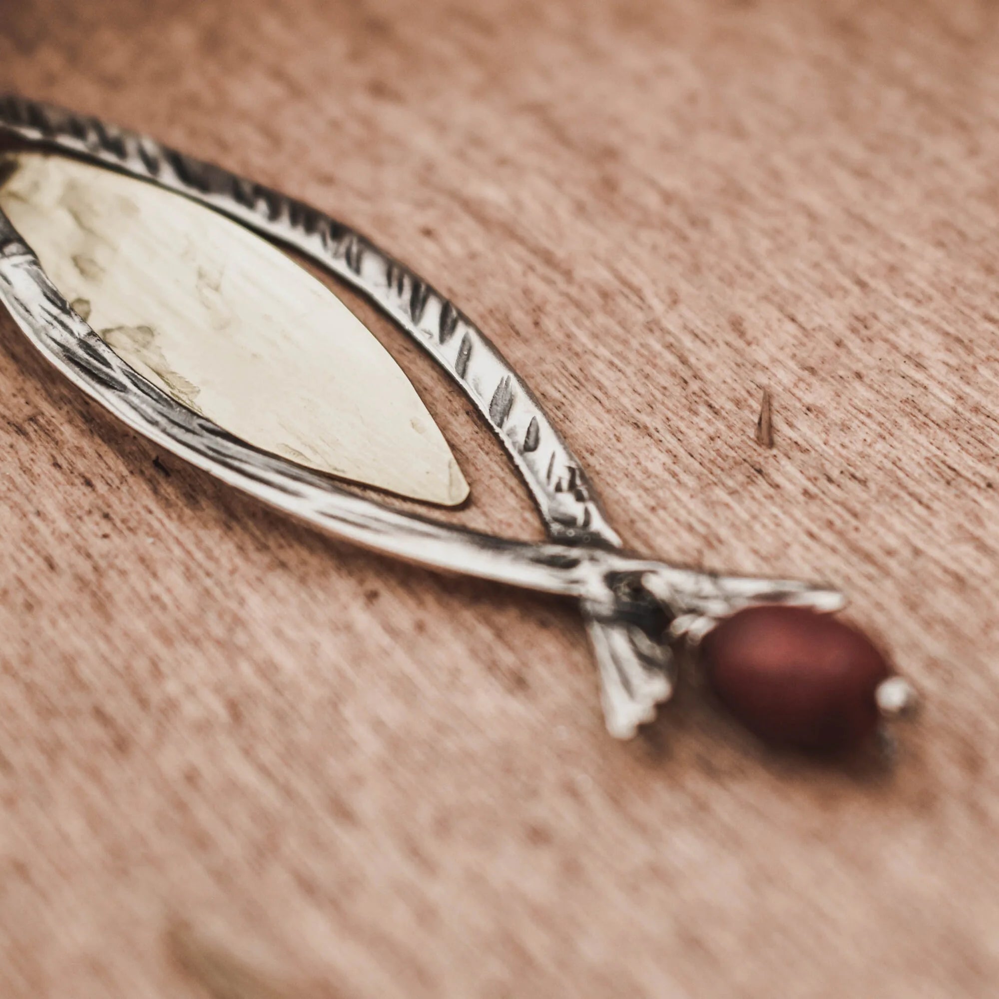 Close-up of a long fish pendant showing the engraved hatched silver frame, smooth gold-plated brass fish body and forked tail with a warm red coral bead drop on a wooden surface.