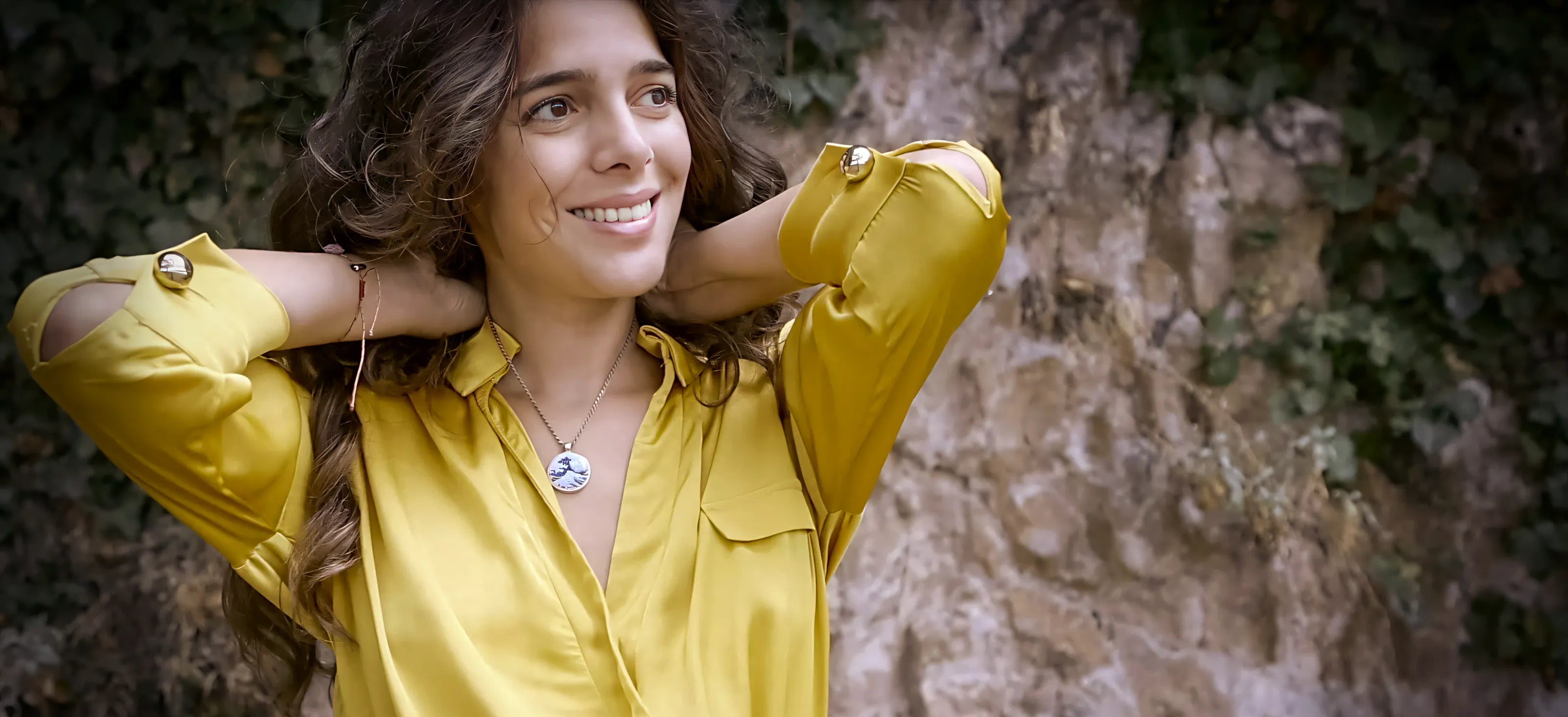 Woman wearing a yellow shirt against a stone wall wearing a ceramic pendant